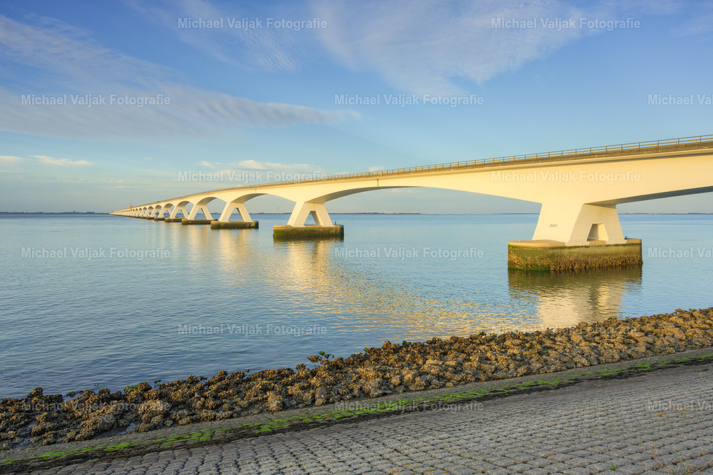 Zeelandbrücke im Licht des frühen Abends | Im warmen Licht des frühen Abends erstreckt sich die imposante Zeelandbrücke über das Wasser und verbindet die Inseln von Zeeland miteinander. Die endlose Reihe der Brückenpfeiler und die klare Linienführung führen den Blick weit in die Landschaft und verleihen dem Motiv eine beeindruckende räumliche Tiefe.Das sanfte Abendlicht taucht die Brücke und die umgebende Küstenlandschaft in eine ruhige, stimmungsvolle Atmosphäre. Ein charakteristisches Landschaftsfoto aus Zeeland, Niederlande, das Architektur und Natur auf eindrucksvolle Weise miteinander verbindet. - Realisiert mit Pictrs.com