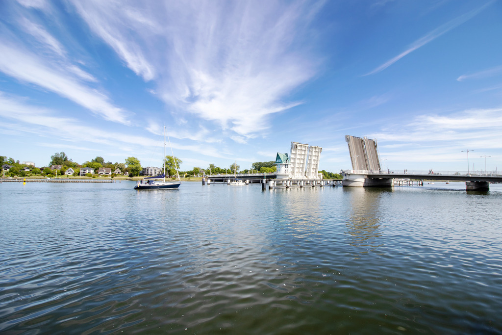Wandbild: Geöffnete Schleibrücke in Kappeln | Dieses Wandbild im Querformat zeigt die geöffnete Schleibrücke in Kappeln an der Schlei. Auf der Schlei befindet sich ein Segelboot. Am blauen Himmel sind einige Schleierwolken zu sehen.  - Realisiert mit Pictrs.com