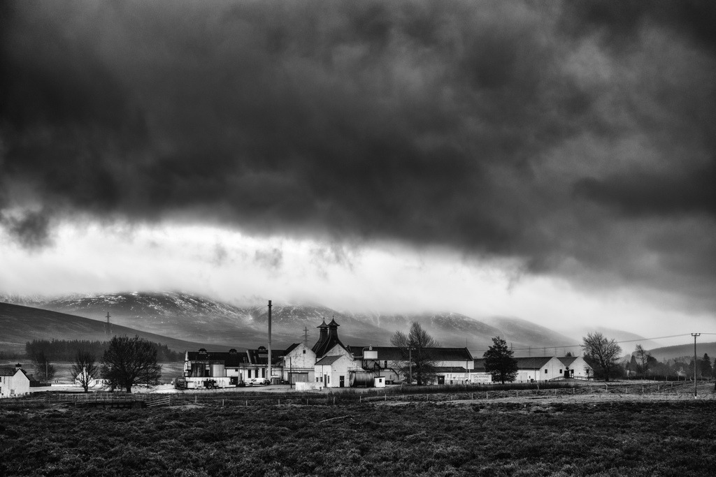 Dalwhinnie Destillerie | Eine Schwarz-Weiß-Aufnahme der Dalwhinnie Destillerie in Schottland zeigt das Gebäudekomplex inmitten einer weiten Landschaft. Dunkle, dramatische Wolken dominieren den oberen Bildbereich, während Nebel die schneebedeckten Berge im Hintergrund umhüllt. Die Szenerie fängt die winterliche Atmosphäre der schottischen Highlands ein.* - Realisiert mit Pictrs.com
