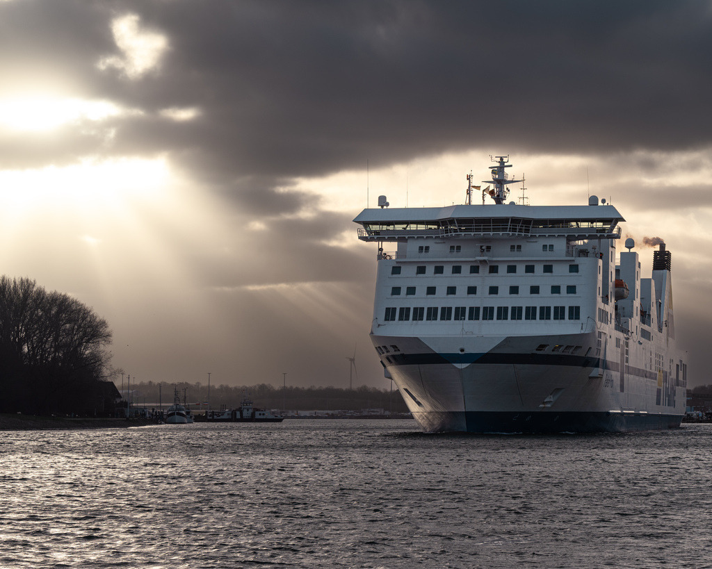 Fähre im Sonnenlicht vor Warnemünde | Eine große Fähre fährt vor Warnemünde durch das aufgewühlte Wasser, während kräftige Sonnenstrahlen durch die dunklen Wolken brechen. Die dramatische Lichtstimmung verleiht der maritimen Szene eine eindrucksvolle Wirkung. - Realisiert mit Pictrs.com