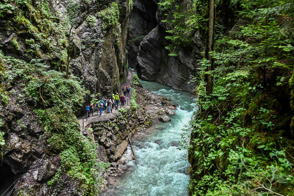 Wildes Naturerlebnis in der Partnachklamm | Spektakuläre Felswände ragen steil empor, während sich das reißende, türkisblaue Wasser der Partnach kraftvoll durch die eng gewundene Schlucht bahnt. Auf dem gesicherten Felsenweg erkunden Wanderer fasziniert dieses naturgewaltige Monument nahe Garmisch-Partenkirchen. Das Zusammenspiel aus rauschendem Wildwasser, moosbewachsenen Felsen und erfrischender Gischt macht die Wanderung durch die Klamm zu einem unvergesslichen Sommerabenteuer, das die ursprüngliche Kraft der Alpen hautnah spürbar macht. - Realisiert mit Pictrs.com