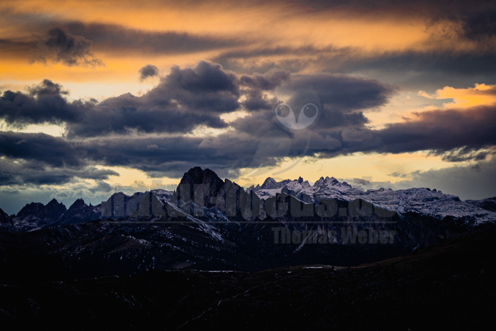 R6NF7254_20251005 | Das Bild zeigt eine weite Ansicht der Dolomiten während des Sonnenuntergangs oder der Dämmerung. Der Himmel ist in leuchtenden Orange- und Gelbtönen gehalten, durchzogen von dunklen, dramatischen Wolkenbändern. Die schroffen, felsigen Gipfel der Berge sind teilweise mit Schnee bedeckt und heben sich dunkel gegen den hellen Himmel ab. Der Vordergrund ist dunkel und undeutlich, zeigt aber die Konturen von Hügeln oder niedrigeren Bergketten. Die Gesamtstimmung ist majestätisch und etwas melancholisch. - Realisiert mit Pictrs.com