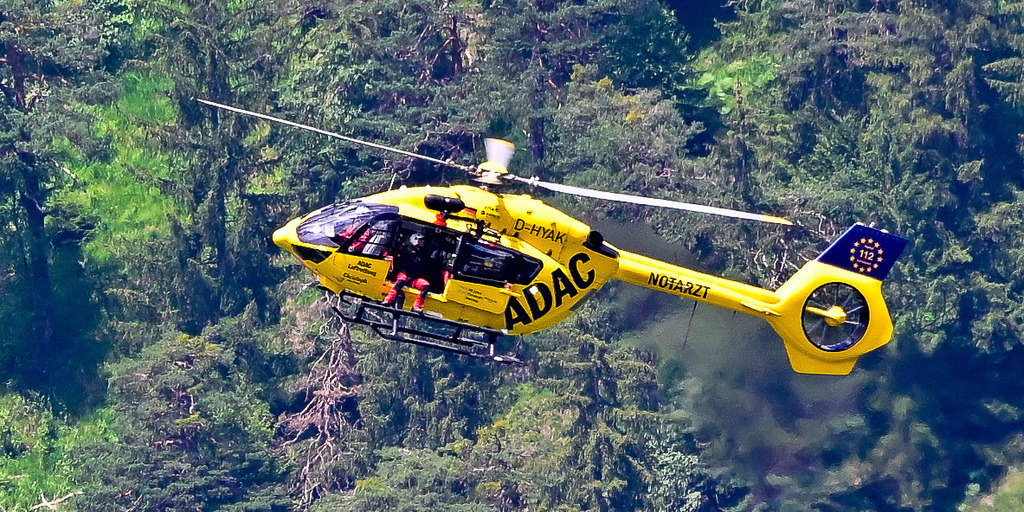 GAP - Juni 2024 | Ein Bild, das pure Dynamik, Erleichterung und tiefes Vertrauen ausstrahlt: Im eleganten Vorwärtsflug zieht der gelbe ADAC-Rettungshubschrauber (D-HYAK) seine Bahn vor einer Steilwand aus dichten, saftig grünen Nadelbäumen. Mit offener Schiebetür, aus der ein Besatzungsmitglied wachsam nach unten blickt, symbolisiert dieser Moment die unermüdliche Einsatzbereitschaft der Bergretter und Notärzte im Hochgebirge. Vor der gewaltigen Kulisse des Bergwaldes wird die Maschine zum leuchtenden Zeichen der Rettung in höchster Not – ein kraftvolles Zeugnis menschlicher Hilfsbereitschaft und modernster Technik, die Leben rettet. - Realisiert mit Pictrs.com