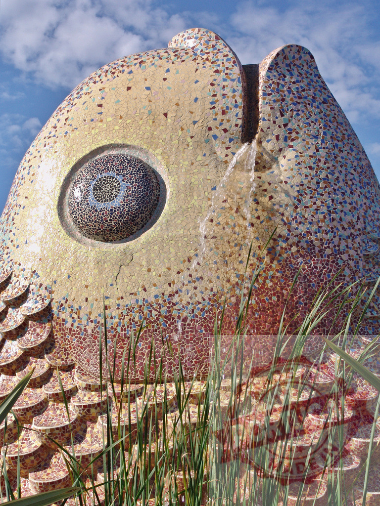 Mosaic fish fountain spouting water against blue sky | Mosaic fish sculpture creating a fountain with water flowing from its mouth - Realized with Pictrs.com