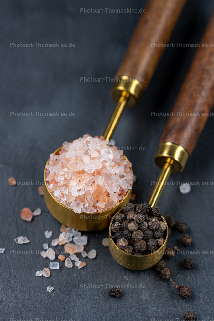 Elegante Gewürz-Messlöffel mit  Salz und Pfeffer | Auf einer edlen Schieferplatte arrangieren sich zwei Messlöffel mit Holzgriffen und goldenen Schalen. Rosa Himalaya-Salz füllt den großen Löffel, schwarzer Pfeffer den kleineren. Die groben Salzkristalle bilden einen reizvollen Kontrast zu den dunklen Pfefferkörnern. Diese geschmackvolle Inszenierung zeigt die Schönheit natürlicher Gewürze und hochwertiger Küchenutensilien in perfekter Harmonie. - Realisiert mit Pictrs.com
