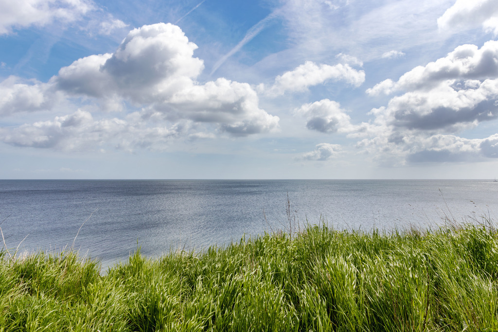 Wandbild: Steilküste an der Ostsee in Schönhagen | Dieses Wandbild im Querformat zeigt den Blick auf die Ostsee von der Steilküste in Schönhagen. Im Vordergrund ist Gras auf der Steilküste zu sehen. Dahinter folgt der Blick auf die offene Ostsee. Am Himmel blauen Himmel befinden sich frühlingshafte helle Wolken. Dieses maritime Wandbild ist in vielen Abmessungen erhältlich. Strandfeeling fürs Zuhause. Ideal fürs Wohnzimmer, Schlafzimmer, Küche, das Büro oder die Praxis.  - Realisiert mit Pictrs.com