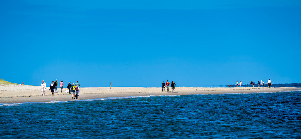 Spaziergänger am Sandstrand der Westfriesischen Insel Texel bei strahlendem Sonnenschein | Eine Gruppe von Urlaubern und Spaziergängern flaniert an einem sonnigen Tag entlang einer weitläufigen Sandbank auf der niederländischen Nordseeinsel Texel. Die Aufnahme zeigt die charakteristische Küstenlandschaft mit feinem Sandstrand, sanftem Wellengang des tiefblauen Meeres und klarem, blauem Himmel. Das Motiv eignet sich ideal für redaktionelle Beiträge zu den Themen Tourismus, Inselurlaub in den Niederlanden, Nordsee, Sommerurlaub, Erholung sowie Natur- und Küstenschutz. - Realisiert mit Pictrs.com