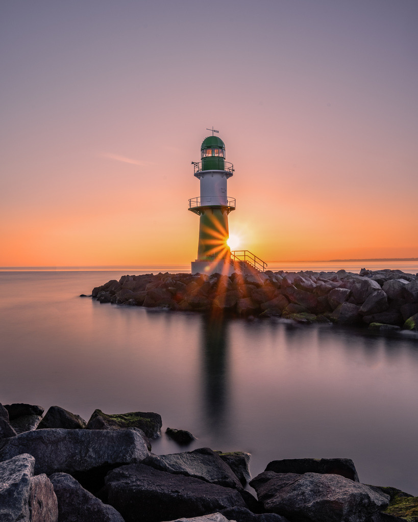 Westmolenfeuer Warnemünde bei Sonnenaufgang | Das grüne Westmolenfeuer von Warnemünde steht während des Sonnenuaufgangs auf der steinernen Mole. Durch die Langzeitbelichtung erscheint die Ostsee besonders ruhig und unterstreicht die klare, minimalistische Komposition. - Realisiert mit Pictrs.com