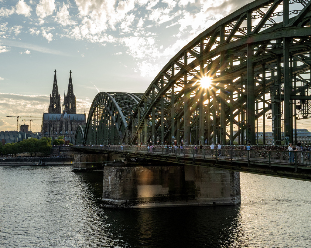 Kölner Dom hinter der Hohenzollernbrücke | Die Hohenzollernbrücke führt den Blick direkt zum Kölner Dom, während die tief stehende Sonne zwischen den Stahlbögen hindurchscheint. Die warme Lichtstimmung verbindet Architektur und Rhein zu einer klassischen Köln-Ansicht. - Realisiert mit Pictrs.com