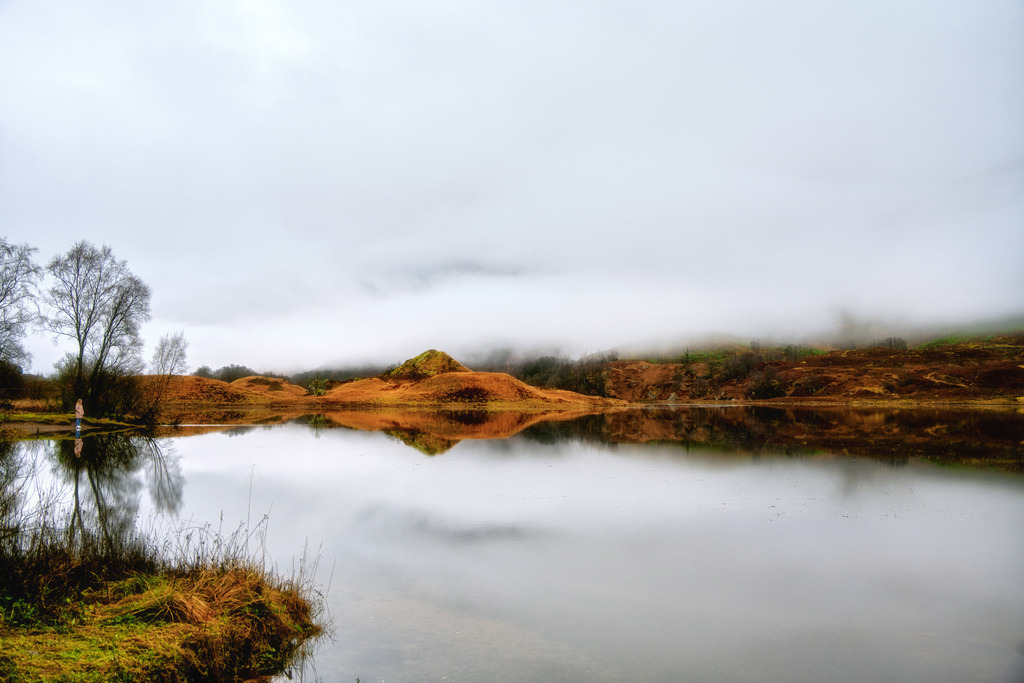 Loch Iubhair im Nebel | Loch Iubhair ist ein idyllischer Süsswassersee im schottischen Glen Dochart, gelegen innerhalb des Loch Lomond and The Trossachs Nationalparks etwa sechs Kilometer östlich von Crianlarich. Der Name bedeutet übersetzt „See der Eiben“. Er ist eng mit dem benachbarten Loch Dochart verbunden und wird vom gleichnamigen River Dochart gespeist. Umgeben von einer beeindruckenden Bergkulisse, insbesondere dem Sgiath Chùil, gilt der See als ruhiger Rückzugsort für Wanderer und Naturliebhaber, die die malerischen Uferabschnitte abseits der grossen Touristenströme schätzen.Eine weite Ansicht des Loch Iubhair in Schottland zeigt die ruhige Wasseroberfläche, die die neblige Landschaft und die kahlen Bäume widerspiegelt. Die umliegenden Hügel sind mit herbstlicher, rötlich-brauner Vegetation bedeckt, während dichter Nebel die oberen Bereiche der Berge verhüllt. Eine einzelne Person am Ufer verleiht der Szene eine menschliche Dimension und betont die Weite der Natur. - Realisiert mit Pictrs.com