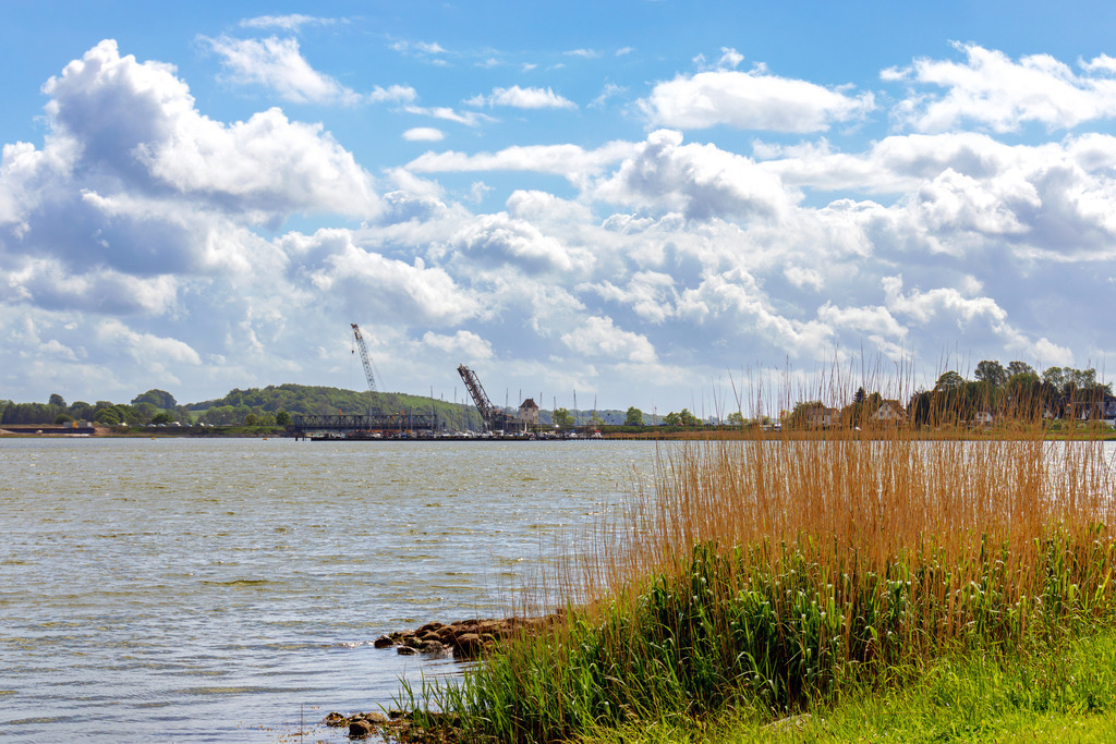 Wandbild: Schleiufer in Lindaunis im Frühling | Dieses Wandbild im Querformat zeigt den Uferbereich an der Schlei im Frühling. In der Ferne ist die Lindaunisbrücke zu sehen. Am blauen Himmel befinden sich einige helle Wolken.  - Realisiert mit Pictrs.com
