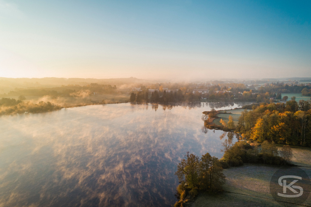 Herbstlandschaft mit Nebel über See - Luftaufnahme Bayern im Morgenlicht | Spektakuläre Drohnenaufnahme einer herbstlichen Seelandschaft im warmen Morgenlicht. Mystischer Bodennebel über dem Wasser, goldene Herbstbäume und verschneite Alpen im Hintergrund vereinen sich zu einem malerischen Panorama in Süddeutschland. - Realisiert mit Pictrs.com