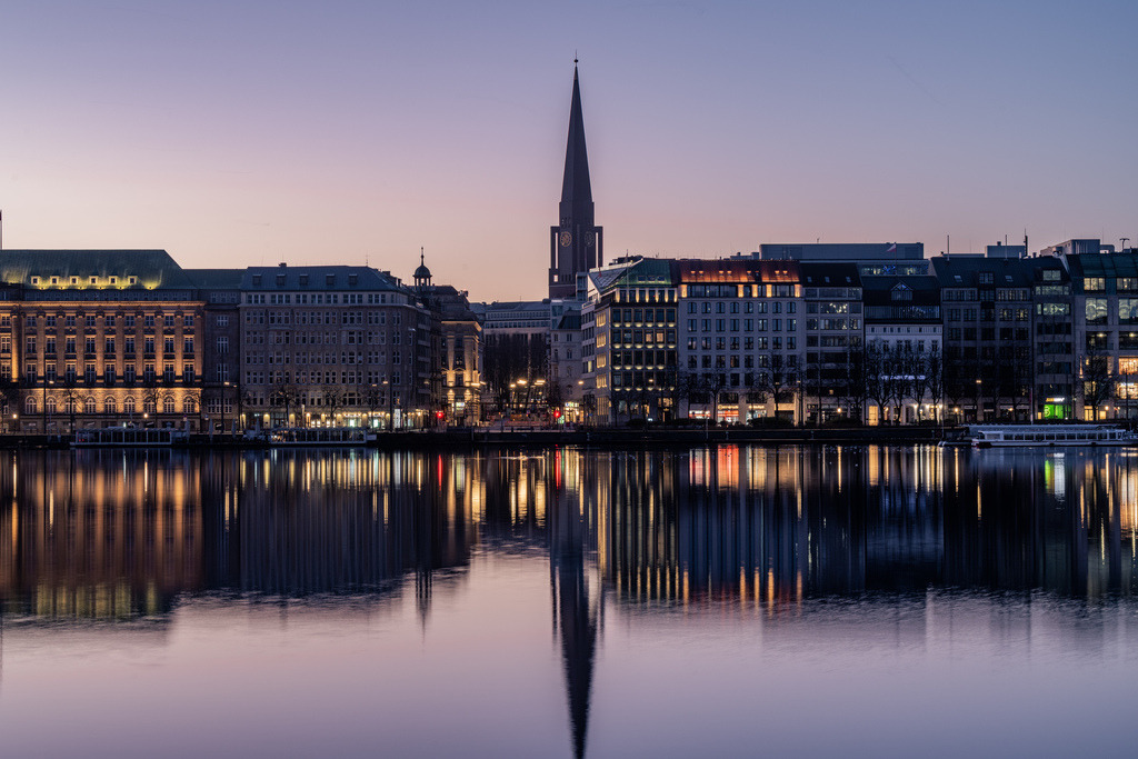 Hamburger Innenstadt an der Binnenalster | Die beleuchteten Fassaden der Hamburger Innenstadt spiegeln sich zur blauen Stunde im ruhigen Wasser der Binnenalster. Der markante Kirchturm und die warmen Lichter prägen die klassische Stadtsilhouette. - Realisiert mit Pictrs.com