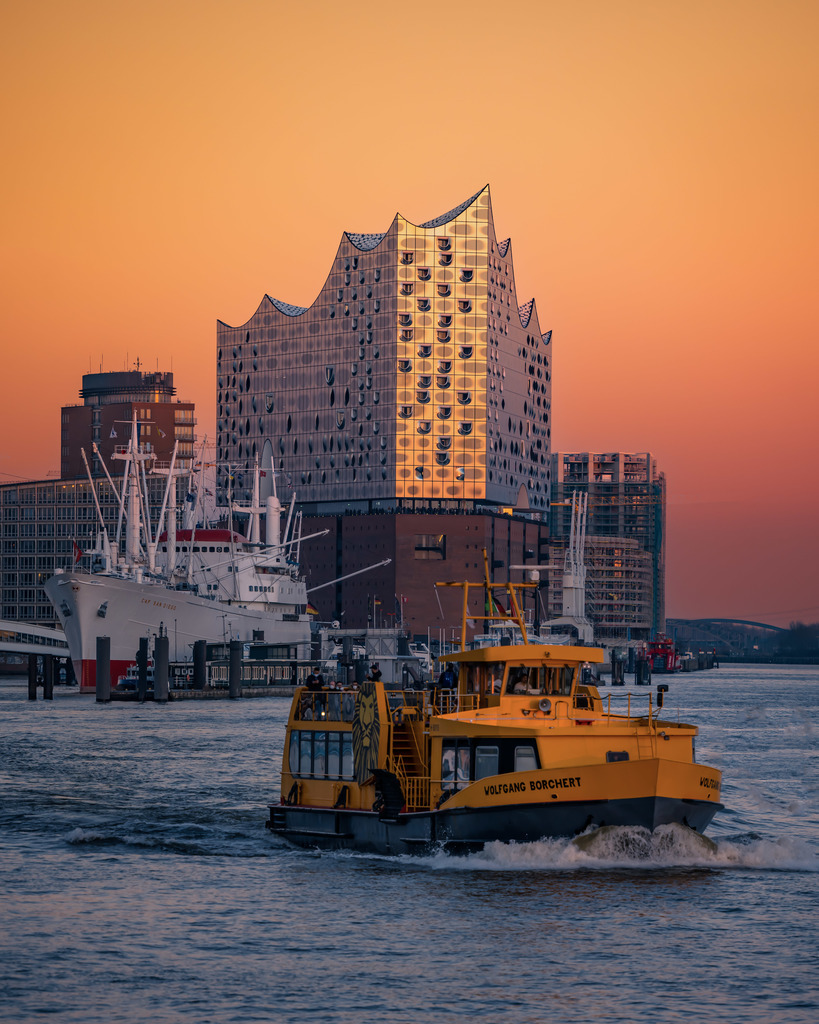 Arbeitsschiff vor der Elbphilharmonie | Ein Arbeitsschiff auf der Elbe vor der Elbphilharmonie im warmen Abendlicht – Hamburger Hafenalltag trifft auf moderne Architektur. - Realisiert mit Pictrs.com