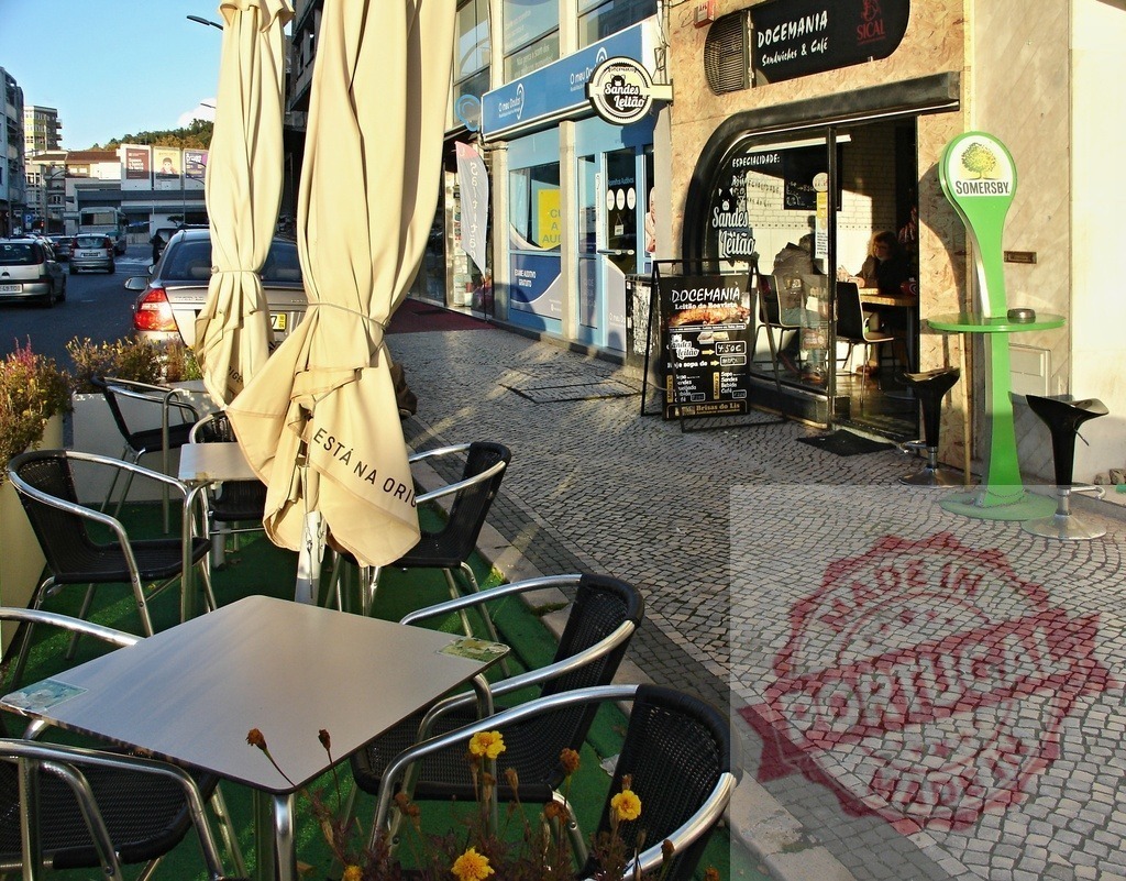 Docemania cafe outdoor terrace on cobblestone street in Portugal | Docemania cafe's outdoor seating area with tables, chairs, and umbrellas on a sunlit cobblestone street - Realisiert mit Pictrs.com