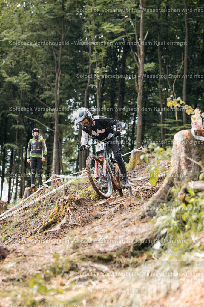 Mountainbike-Abenteuer im Wald | Das Foto zeigt einen Mountainbiker in voller Montur, der einen steilen, erdigen Hang hinunterfährt. Er trägt einen schwarzen Helm mit einer weißen Nummer '65' und eine dunkle Brille. Seine Kleidung ist hauptsächlich schwarz und weiß. Im Hintergrund ist ein weiterer Mountainbiker in ähnlicher Ausrüstung zu sehen. Der Hang ist mit Ästen und Blättern bedeckt, und der Wald ist dicht und grün. Das Wetter scheint bewölkt zu sein. - Realisiert mit Pictrs.com