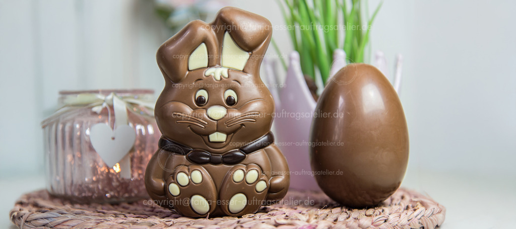 Schokoladen Hase und Ei D | Ein Schokoladen Hase und ein Schokoladen Ei stehen auf einem Tisch, der mit Hyazinthen und Kerzen dekoriert ist. Verschwommene pinke Deko Elemente. 