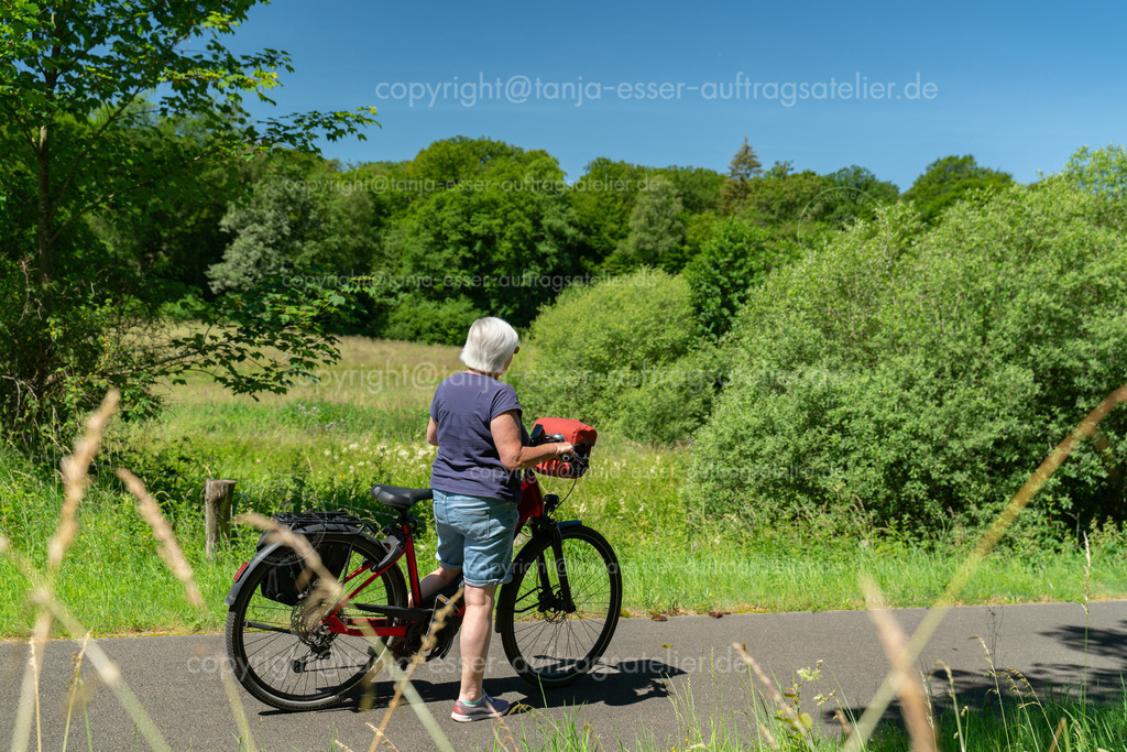 Ältere Frau mit E Bike auf dem Möhnetal Radweg schaut in die Auenlandschaft | Eine ältere Frau mit einem roten modernen E Bike steht am Möhnetal Radweg in Brilon und blickt in die Auenlandschaft mit saftigen Wiesen entlang dem Fluss Möhne im Sauerland.