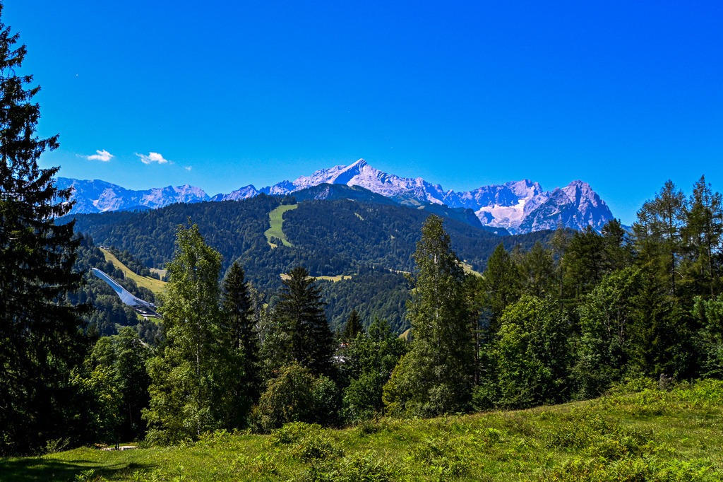 Weitblick über Garmisch-Partenkirchen | Ein faszinierendes Panorama fängt die ganze Vielfalt des Werdenfelser Landes ein. Unter einem strahlend blauen Himmel erstreckt sich das gewaltige Zugspitzmassiv mit der markanten Alpspitze. Im grünen Tal blitzt links die ikonische Olympiaschanze von Garmisch-Partenkirchen zwischen den Bäumen auf. Dichte Mischwälder und saftige Bergwiesen im Vordergrund rahmen die Szene perfekt ein. Ein Bild, das die harmonische Verbindung von alpiner Sporttradition und unberührter Natur spürbar macht. - Realisiert mit Pictrs.com