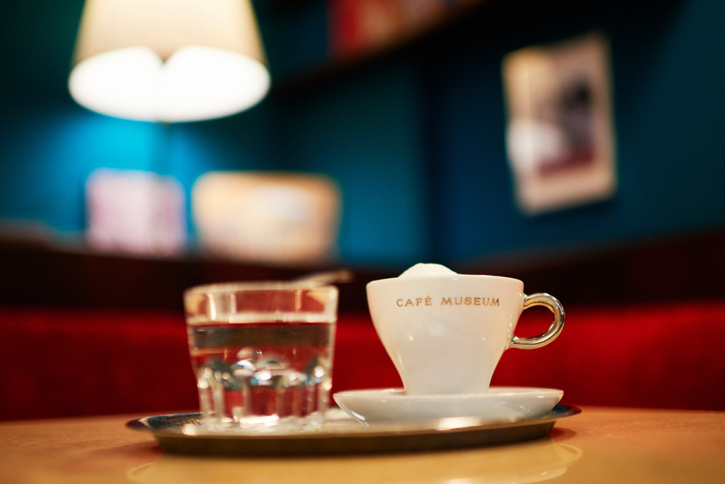 Cafe Museum | Wien, Austria - June 22, 2018: Café Museum, eine Tasse Melange und ein Glas Wasser auf dem Tisch stehend, im Hintergrund Bücher und Bilder. - Realisiert mit Pictrs.com