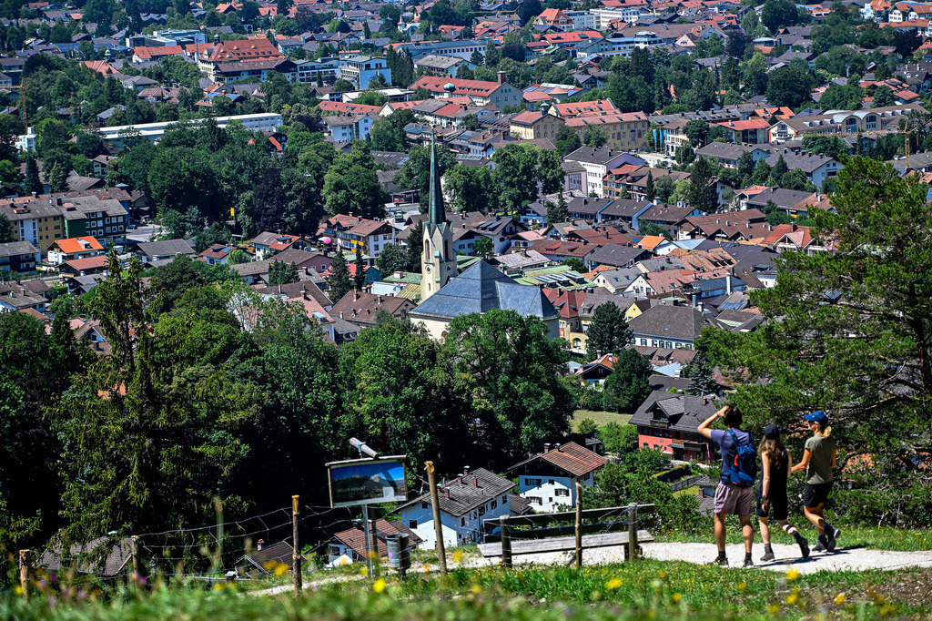 Der Blick auf Partenkirchen nach dem Aufstieg | Ein malerischer Ausblick über den Ortsteil Partenkirchen belohnt die Mühen des Aufstiegs: Drei Wanderer spazieren im warmen Sonnenlicht einen Bergpfad entlang, während im Hintergrund die charmanten Dächer des Ortes und die markante Kirchturmspitze der Pfarrkirche Maria Himmelfahrt emporragen. Eingerahmt von saftigem Grün und schattigen Bäumen fängt dieses Motiv das Gefühl von Freiheit, bayerischer Gemütlichkeit und der Sehnsucht nach alpiner Natur perfekt ein. - Realisiert mit Pictrs.com