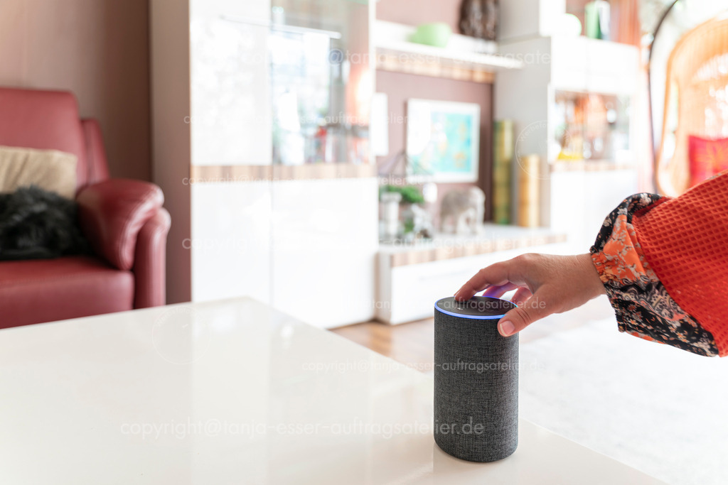 Smarter Lautsprecher im Wohnzimmer weibliche Hand D | Ein smarter Lautsprecher Assistent steht in einem hellen Wohnzimmer. Eine weibliche Hand betätigt eine Taste. Viel Copy Space.
