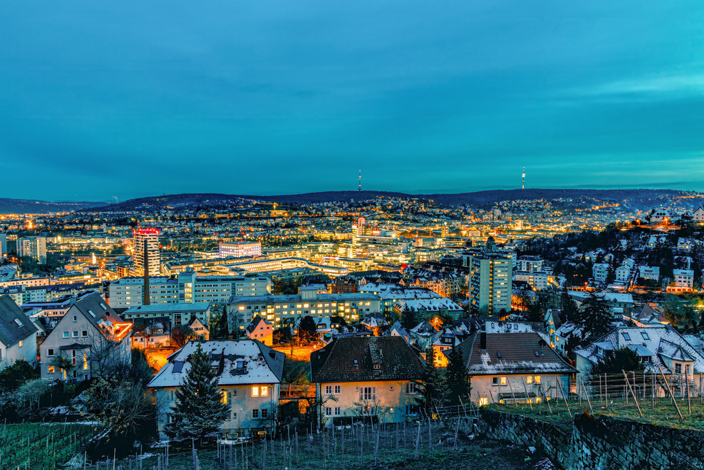 Nächtlicher Panorama-Blick im Winter über die Stadt Stuttgart | Das Bild zeigt einen nächtlichen Panorama-Blick über die Stadt Stuttgart. Es handelt sich um die baden-württembergische Landeshauptstadt, bekannt als "Schwabenmetropole". Typisch für Stuttgart ist die Kessellage, die auf dem Foto durch die umliegenden Höhenzüge angedeutet wird. Die Lichter der Stadt beleuchten das dichte Stadtgebiet, während der Himmel in den frühen Abendstunden oft noch ein tiefes Blau zeigt.  - Realisiert mit Pictrs.com