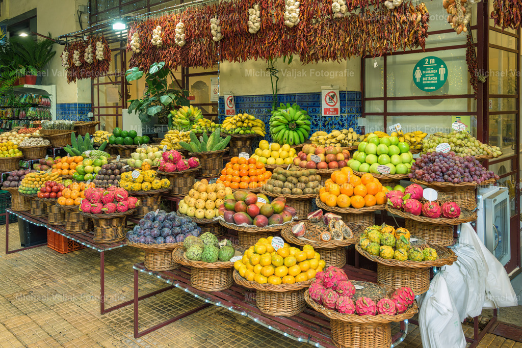 Obststand in der Markthalle in Funchal auf Madeira | Ein farbenfroher Obststand in der Markthalle von Funchal ist ein Anblick, der alle Sinne anspricht. Auf diesem Foto sieht man eine Vielzahl von bunten Früchten, die in perfekten Stapeln arrangiert sind.Die Markthalle selbst, auch bekannt als Mercado dos Lavradores, ist ein historisches Bauwerk und ein wichtiger Bestandteil des kulturellen Lebens in Funchal, der Hauptstadt von Madeira. Hier treffen sich Einheimische und Touristen, um frische Produkte, lokale Spezialitäten und handwerkliche Erzeugnisse zu kaufen.Ein besonderes Highlight dieses Marktes sind die exotischen Früchte, die auf Madeira angebaut werden. Dazu gehören die Passionsfrucht, die Pitanga und die Ananasbanane, die alle ihren eigenen einzigartigen Geschmack und Charme haben. Das Foto fängt die lebendige Atmosphäre und die Vielfalt des Angebots perfekt ein und lädt dazu ein, den Obststand genauer zu erkunden und die frischen, aromatischen Früchte zu kosten. - Realisiert mit Pictrs.com