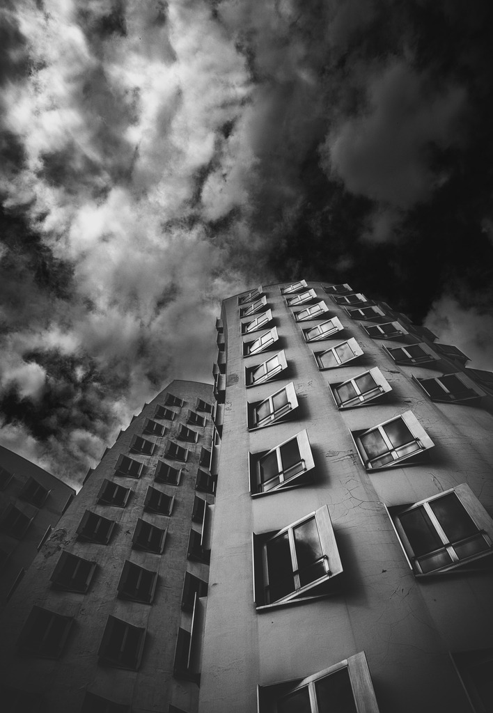 Windows to the Sky | Dramatische Schwarzweißaufnahme moderner Hochhausarchitektur aus extremer Froschperspektive. Die geöffneten Fenster durchbrechen die strenge Geometrie der Fassade und erzeugen einen spannenden Kontrast zwischen Ordnung und Individualität. Über den Gebäuden türmen sich eindrucksvolle Wolkenformationen, die dem Bild eine fast surreale und cineastische Wirkung verleihen.Ein ausdrucksstarkes Fine-Art-Motiv für Liebhaber von Urban Photography, Architektur und atmosphärischen Schwarzweißkompositionen. - Realisiert mit Pictrs.com