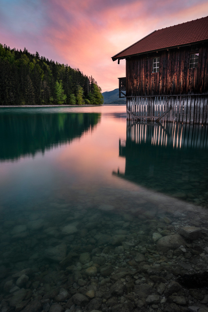 Abendruhe am Walchensee | Sanfte Farben spiegeln sich im glasklaren Wasser, die Linien der Hütte führen den Blick in die Tiefe der Szene. Der Walchensee zeigt sich in diesen Momenten von seiner stillsten Seite. - Realisiert mit Pictrs.com