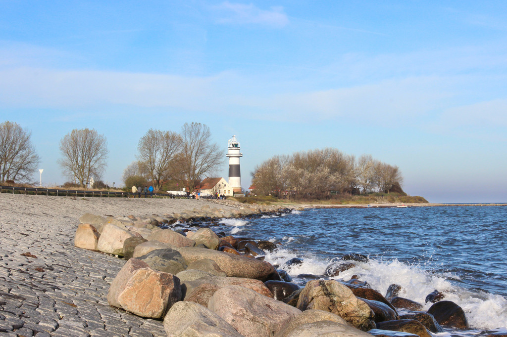 Leuchtturm in Bülk | Dieses Wandbild im Querformat zeigt den Leuchtturm an der Kieler Förde in Bülk in der Ferne. Im Vordergrund ist der Uferbereich sowie einige kleine Wellen zu sehen. Am blauen Himmel befinden sich nur wenige kleine Wolken.  - Realisiert mit Pictrs.com