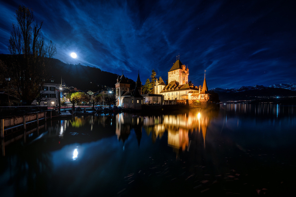 Schloss Oberhofen bei Mondlicht | Das Schloss Oberhofen am Thunersee ist bei Nacht beleuchtet und spiegelt sich im ruhigen Wasser. Ein heller Mond und langzeitbelichtete Wolken dominieren den tiefblauen Nachthimmel über den schneebedeckten Bergen im Hintergrund. Die Aufnahme betont die malerische Architektur des Schlosses und die friedliche Atmosphäre der alpinen Seenlandschaft. - Realisiert mit Pictrs.com