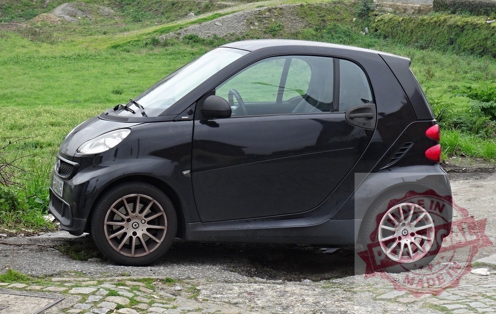 Black Smart car parked outdoors on paved area | Black Smart car parked on pavement next to a green grassy slope - Realisiert mit Pictrs.com