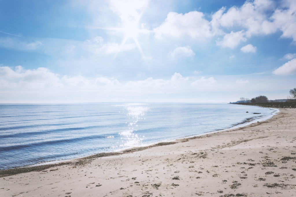 Sonnenlicht über der Ostsee – Heller Strandbogen in Damp | Dieses maritime Wandbild aus Damp zeigt einen Strandabschnitt, in dem das Sonnenlicht über dem Wasser glitzert und die Wellen in feinen Reflexen aufleuchten. Der sanft geschwungene Küstenverlauf und die hellen Wolken verleihen dem Motiv eine offene, lichtbetonte Atmosphäre. Die Aufnahme fängt den Moment ein, in dem Licht, Wasser und Strand eine klare, freundliche Küstenszene formen.