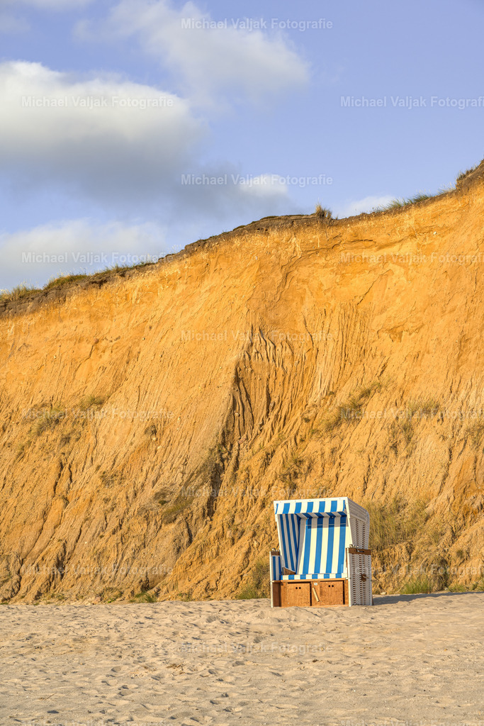 Strandkorb am Roten Kliff auf Sylt | Ein einzelner Strandkorb steht vor der Abbruchkante des Roten Kliffs bei Kampen auf Sylt. Die Abendsonne taucht die Steilküste in ein orangefarbenes Licht.  - Realisiert mit Pictrs.com