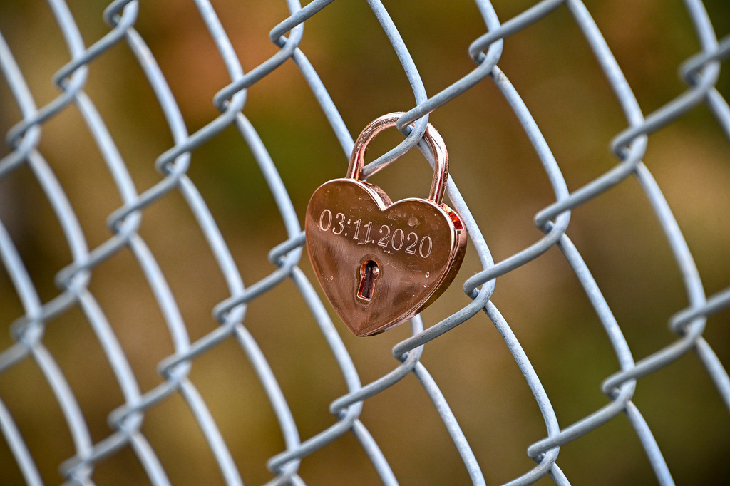 Für immer wir: Ein Versprechen in Roségold | An einem schlichten Maschendrahtzaun hängt es – klein, glänzend und voller Bedeutung. Ein herzförmiges Vorhängeschloss in warmem Roségold, das den 3. November 2020 für die Ewigkeit festhält. Während die Zeit unaufhaltsam verstreicht und die Welt im Hintergrund in sanften, herbstlichen Farben verschwimmt, bleibt dieses kleine Schloss ein unerschütterliches Symbol für ein stilles Versprechen. Es erzählt die Geschichte zweier Menschen, die an diesem Tag Ja zueinander gesagt haben – festgehalten für die Unendlichkeit. - Realisiert mit Pictrs.com