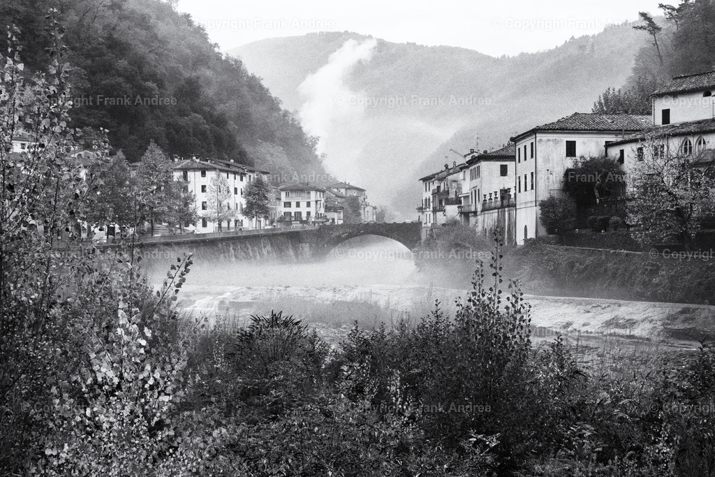 Bagni di Lucca | Schwarz weiß Fotografie von Bagni die Lucca in der Toskana an einem nebligen Tag. Blick über den Fluss Lima, über dem Nebel zieht, zu den Häusern der Altstadt. Im Hintergrund die Berge der Toskana die im Nebel verblassen. - Realisiert mit Pictrs.com