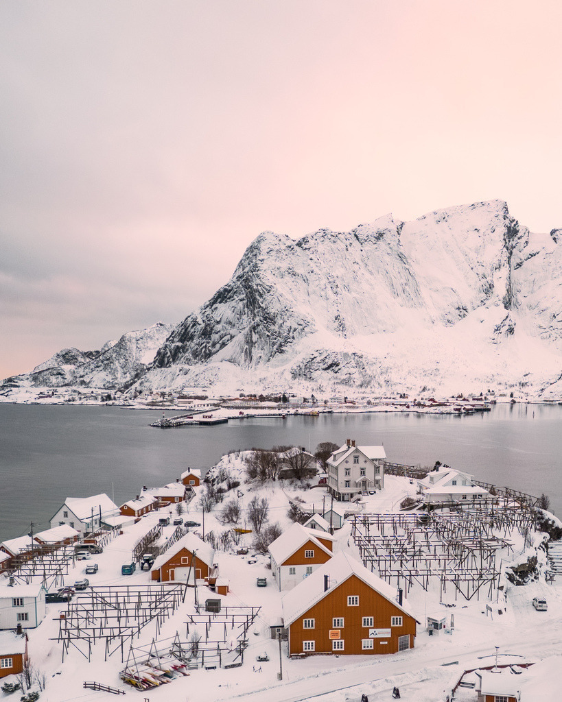 Norwegisches Winterdorf im Morgenlicht | Ein kleiner norwegischer Küstenort liegt unter einem mächtigen verschneiten Berg. Das sanfte rosafarbene Morgenlicht verleiht der winterlichen Landschaft eine ruhige und fast unwirkliche Atmosphäre. - Realisiert mit Pictrs.com