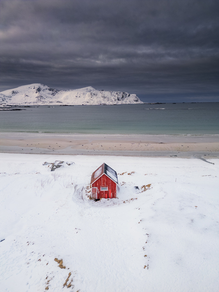 Rote Hütte am verschneiten Strand | Eine einzelne rote Hütte steht inmitten einer weiten, verschneiten Küstenlandschaft. Der starke Farbkontrast zwischen dem Gebäude, dem Schnee und dem dunklen Himmel prägt die minimalistische Komposition. - Realisiert mit Pictrs.com