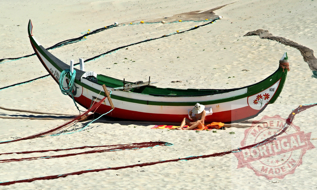 Woman relaxing on beach reading book by fishing boat | Woman in hat reading book on towel beside traditional fishing boat on sandy beach - Realized with Pictrs.com