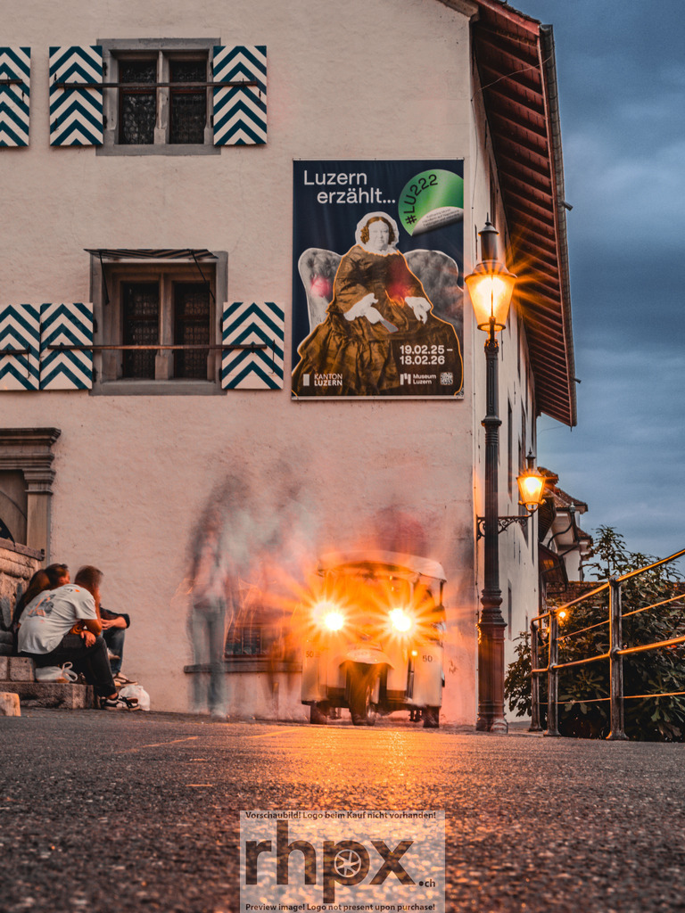 Lichtspiel am Historischen Museum | Licht. Kontrast. Geschichte.Die hellen Lichter eines E-Tuks setzen einen modernen Akzent vor der historischen Fassade des Luzerner Museums. Das warme Leuchten der Strassenlaterne und die kühle Abendstimmung schaffen eine lebendige Atmosphäre in den Gassen der Stadt.Wähle unter "Produktauswahl" dein Wunschformat: Vom klassischen Wandbild über Puzzle &amp; Tassen bis zum digitalen Download (z.B. als Handy-Hintergrund). - Realisiert mit Pictrs.com
