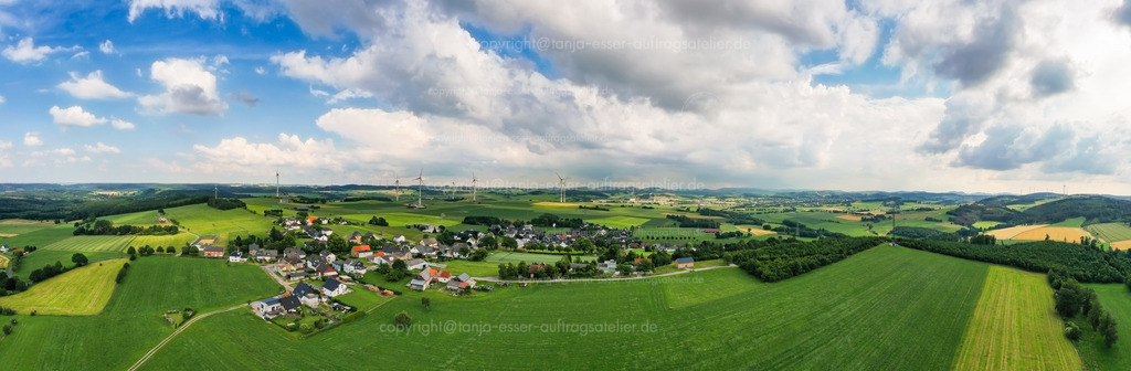 Panoramafoto von Sauerländer Ortschaft Wülfte | Luftbild Panorama des Ortsteil Wülfte bei Brilon im Sauerland. Häuser, Felder, Bauernhöfe und eine Kapelle vor grünen Hügeln. Blauer Himmel mit Sonnenschein.