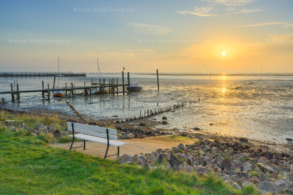 Ein Platz am Wattenmeer | Das Wattenmeer, ein UNESCO-Weltnaturerbe, bietet mit seinem einzigartigen Ökosystem einen atemberaubenden Anblick, besonders während des Sonnenaufgangs. In Steenodde auf Amrum offenbart sich die Schönheit dieses Ortes, wenn das erste Licht des Tages den Hafen erleuchtet und die weiße Bank im Vordergrund zum idealen Platz für eine morgendliche Rast wird. Bei Niedrigwasser entfaltet sich ein weiter Blick über das Watt, ein Moment der Ruhe und der natürlichen Schönheit. - Realisiert mit Pictrs.com