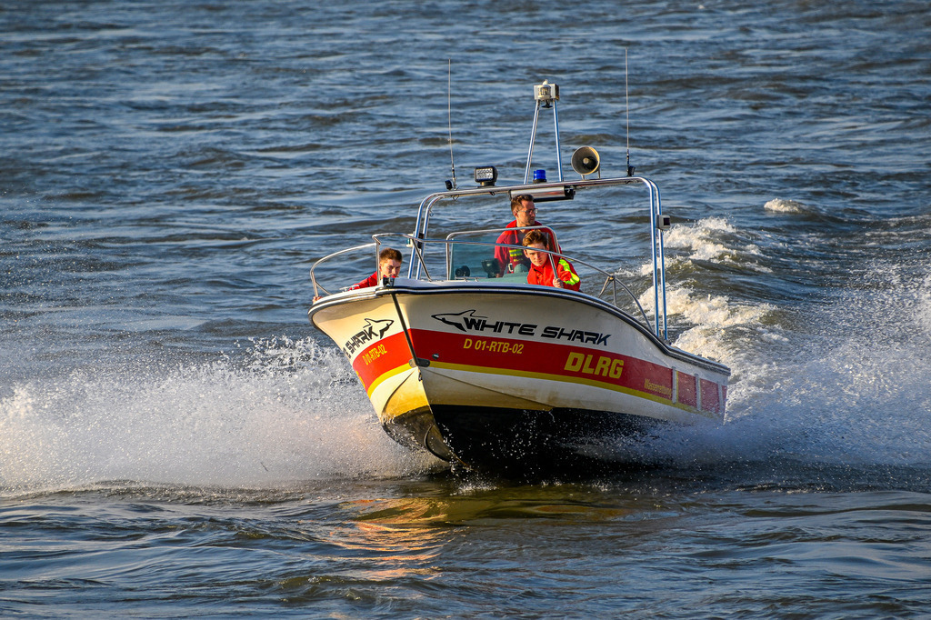 Einsatz auf den Wellen: Die DLRG in Aktion | Das Bild zeigt ein Rettungsboot der DLRG mit dem Namen „White Shark“ (Kennung D 01-RTB-02), das mit hoher Geschwindigkeit über den Rhein bei Düsseldorf fährt. Am Bug und Steuerstand sind drei Einsatzkräfte in ihrer typischen rot-gelben Schutzkleidung zu sehen. Das Boot schneidet dynamisch durch das Wasser, wodurch eine markante Gischt- und Heckwelle entsteht. Das warme Sonnenlicht des späten Nachmittags beleuchtet die Szene und lässt das Wasser in tiefen Blautönen schimmern. - Realisiert mit Pictrs.com