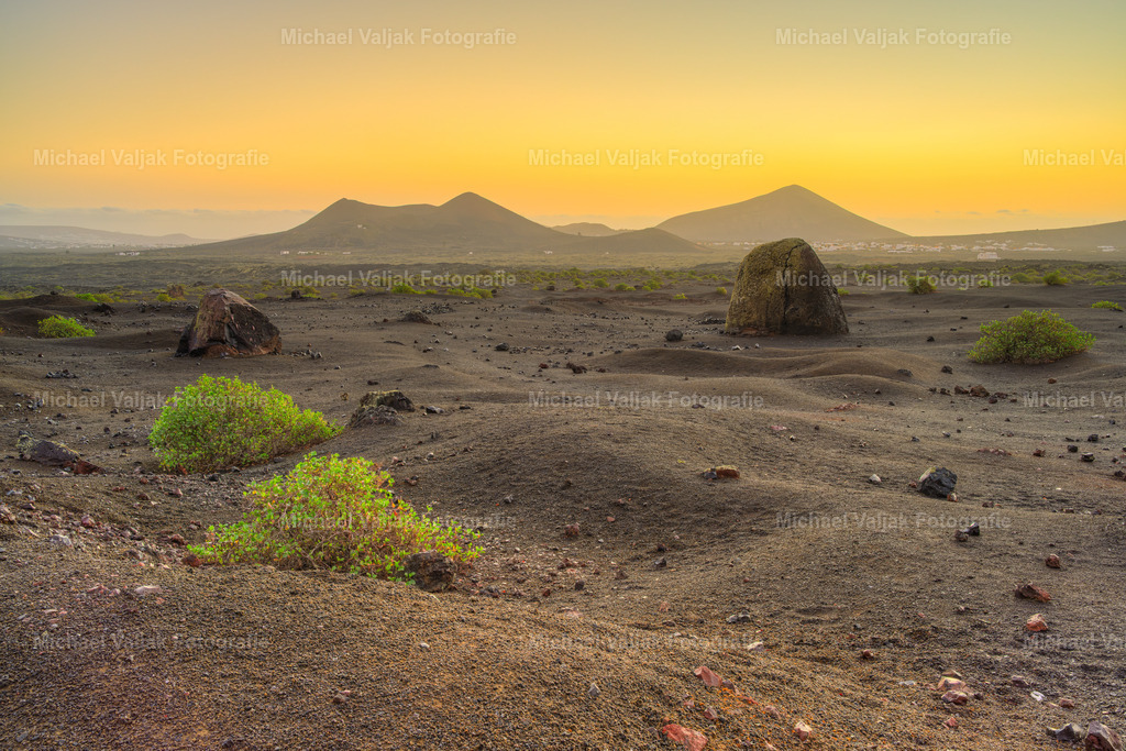 Vulkanlandschaft auf Lanzarote in der Morgendämmerung | Vor dem ersten Tageslicht ruht die gewaltige Vulkanbombe am Montana Colorada wie ein stummer Zeuge vergangener Eruptionen. Dahinter erscheinen der Volcán de Juan Bello, die Häuser von Masdache und der Montana Blanca in sanften Konturen. Mit dem nahenden Sonnenaufgang legt sich ein warmer Schimmer über die Szene – die Farben der Insel beginnen zu leuchten und verleihen der kargen Landschaft eine stille, fast feierliche Schönheit. - Realisiert mit Pictrs.com