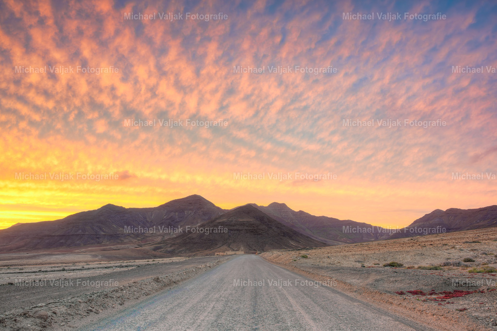 Straße zum Punta de Jandia auf Fuerteventura | Die unbefestigte Straße zum Punta de Jandía zieht sich durch die karge Landschaft und läuft direkt auf die Berge am Horizont zu. Der Sonnenuntergang taucht den Himmel in kräftige Farben, warmes Orange mischt sich mit Rot und Violett. Das Licht legt sich weich über die Piste und die Felsen, als würde der Tag die Insel noch einmal in voller Intensität zeigen. Ein Moment, der die Ruhe, Weite und Rauheit des Südens von Fuerteventura perfekt einfängt. - Realisiert mit Pictrs.com