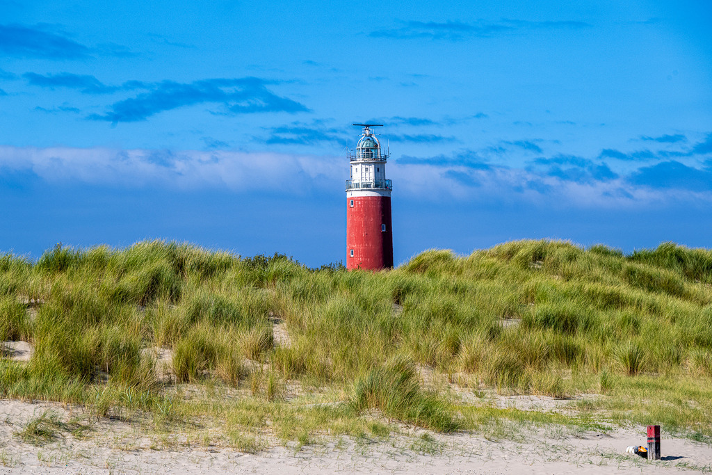 Rot-weißer Leuchtturm Eierland in den Dünen auf der Nordseeinsel Texel | Der charakteristische rote Leuchtturm Eierland ragt an der Nordspitze der niederländischen Insel Texel über die saftig grünen Dünen hinaus. Die Aufnahme zeigt das markante Wahrzeichen bei leicht bewölktem, blauem Himmel. Das Motiv eignet sich ideal für redaktionelle Berichte über den Tourismus in den Niederlanden, Urlaub an der Nordsee, maritime Architektur, Küstenschutz sowie Reiseberichte über die Westfriesischen Inseln. - Realisiert mit Pictrs.com