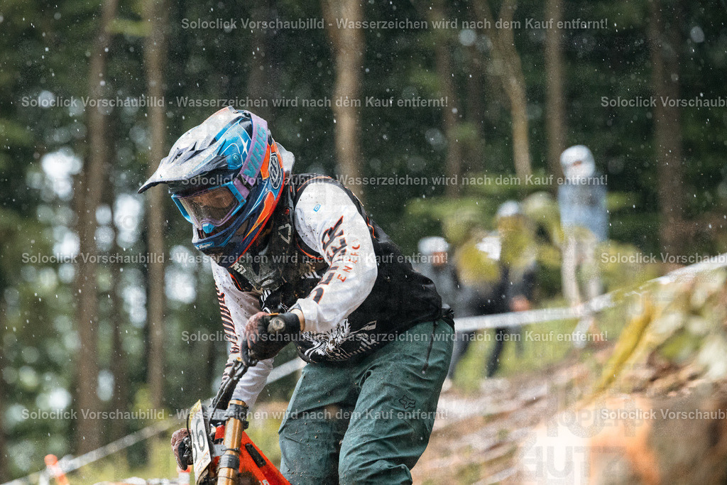 Mountainbiker im Regen | Das Foto zeigt einen Mountainbiker in voller Ausrüstung, der einen steilen, bewaldeten Trail hinunterfährt. Der Biker ist in voller Montur, inklusive Helm, Goggles und Schutzkleidung. Der Trail ist schlammig und steil, und es regnet. Der Hintergrund besteht aus dichtem Grünzeug und Bäumen. - Realisiert mit Pictrs.com