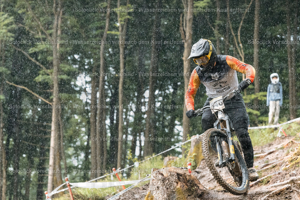 Mountainbiker im Regen | Das Foto zeigt einen Mountainbiker, der einen steilen, bewaldeten Hang im Regen hinabfährt. Der Biker trägt einen Helm und Schutzkleidung und fährt ein Mountainbike mit Federgabel und breiten Reifen. Der Hintergrund besteht aus dichtem Grün und einem roten Band, das den Weg markiert. Es regnet stark, was die Szene dynamisch und herausfordernd erscheinen lässt. - Realisiert mit Pictrs.com