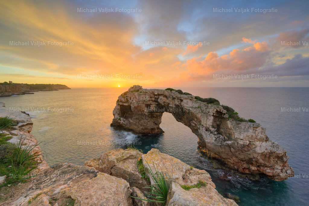 Ein neuer Tag beginnt über Es Pontàs auf Mallorca | Die Sonne hebt sich eben über den Horizont, und ihr warmes Licht taucht Es Pontàs in ein sanftes Glühen. Der Felsbogen zeichnet sich klar vor dem noch farbintensiven Himmel ab, während das Meer in Gold und Orange aufleuchtet. Ein Augenblick, in dem der neue Tag spürbar beginnt. - Realisiert mit Pictrs.com